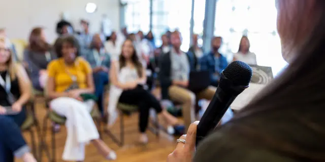 Confident female business conference speaker addresses a large group of business people.