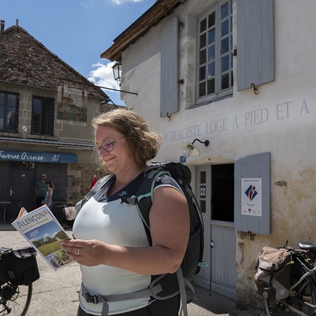 Randonneuse feuilletant le guide touristique d'Alençon Tourisme devant l'Office de tourisme de Saint-Céneri-le-Gérei