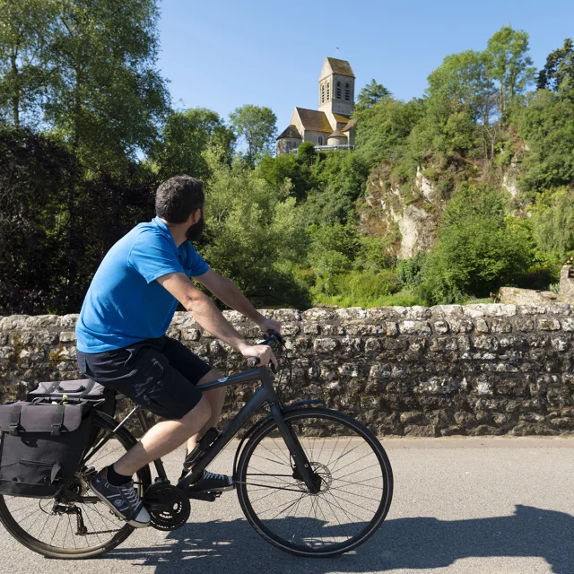 Un cycliste traversant le pont en pierre de Saint-Céneri-le-Gérei et regardant l'église romane