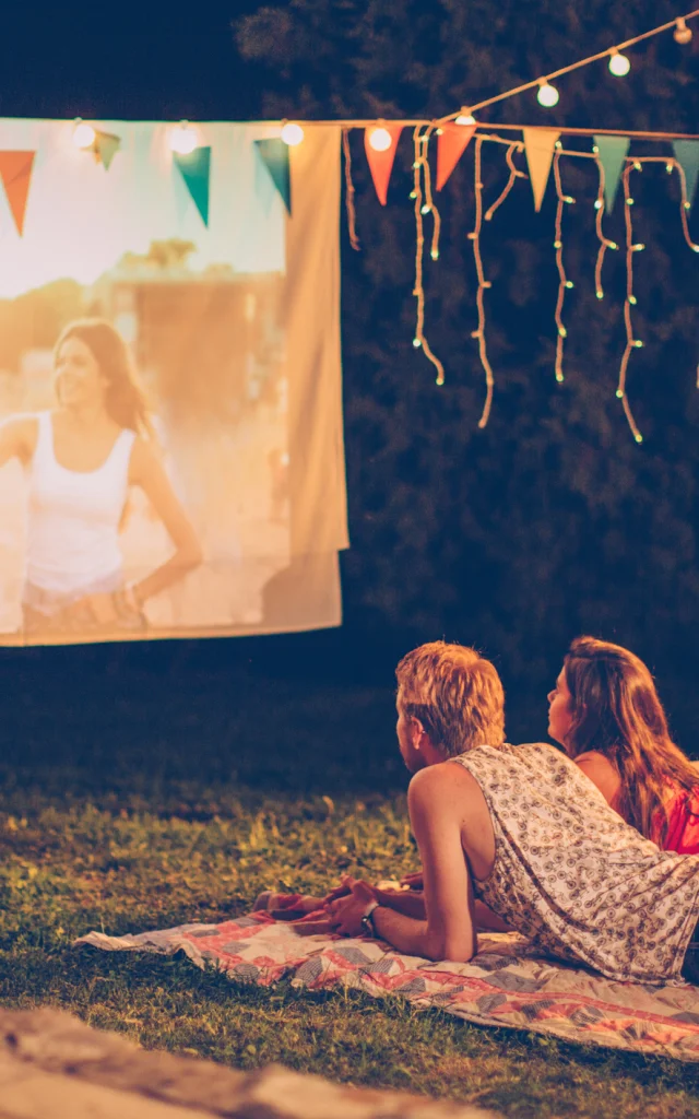 Young couple having movie night party. Laying down on blanket in front of movie improvised screen. Backyard decorated with festive string lights. Night time.