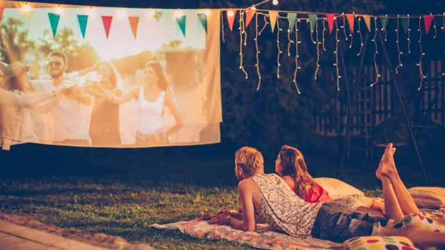 Young couple having movie night party. Laying down on blanket in front of movie improvised screen. Backyard decorated with festive string lights. Night time.