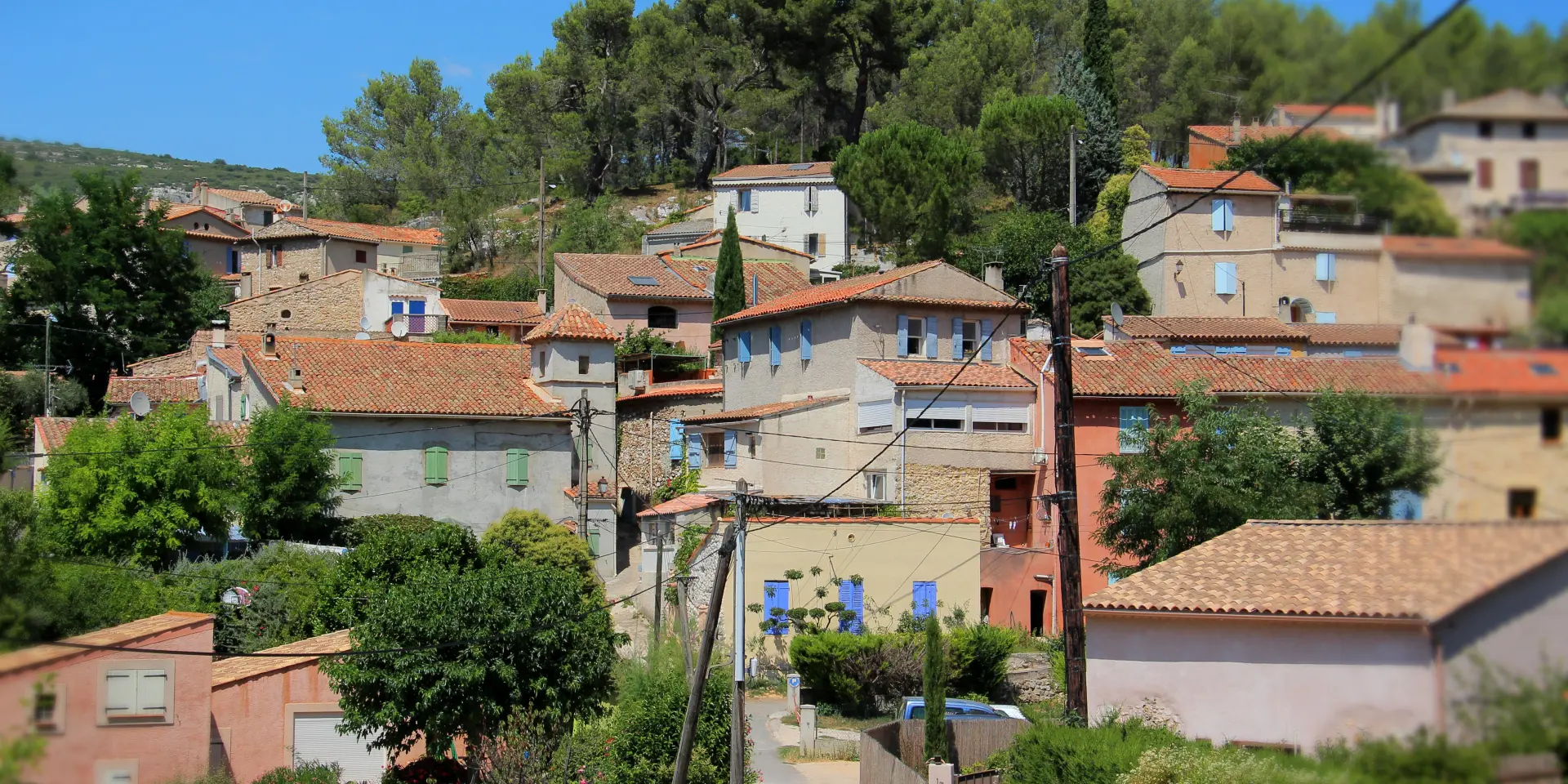 Vue d’ensemble d’un village provençal avec maisons en pierre et façades claires, toits en tuiles rouges, volets colorés, rue en pente et collines boisées sous un ciel bleu à la Bouilladisse