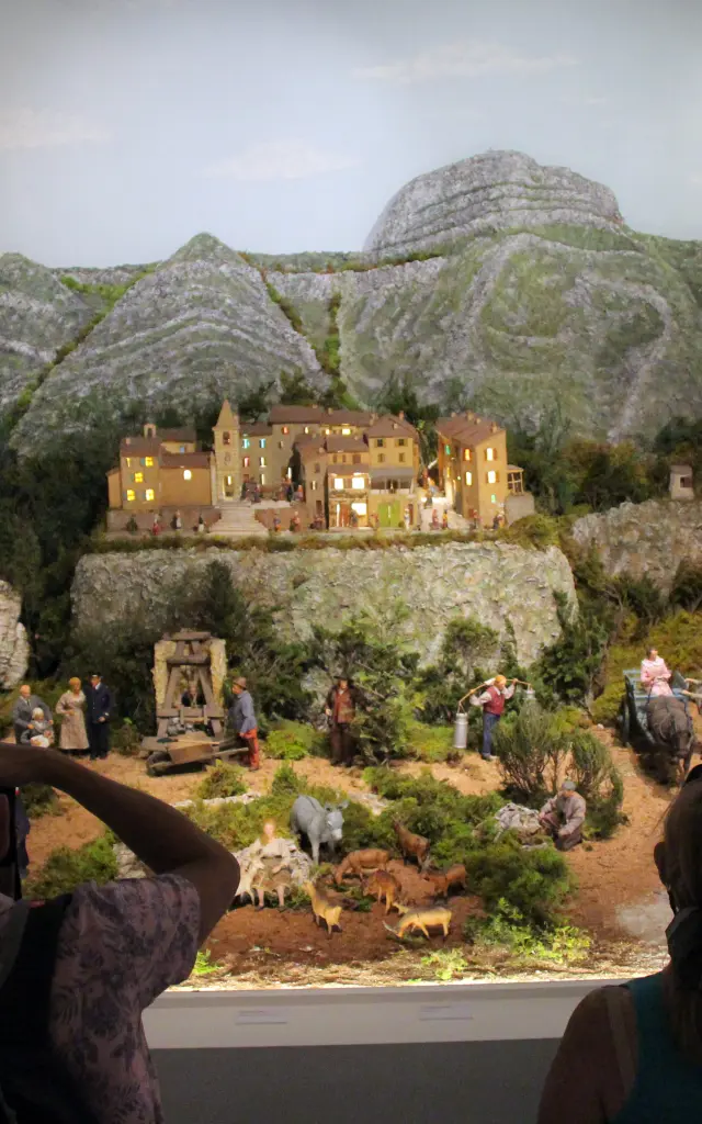 Diorama de village provençal avec maisons miniatures, collines rocheuses et personnages, observé par des visiteurs en intérieur au Petit Monde de Marcel Pagnol à Aubagne