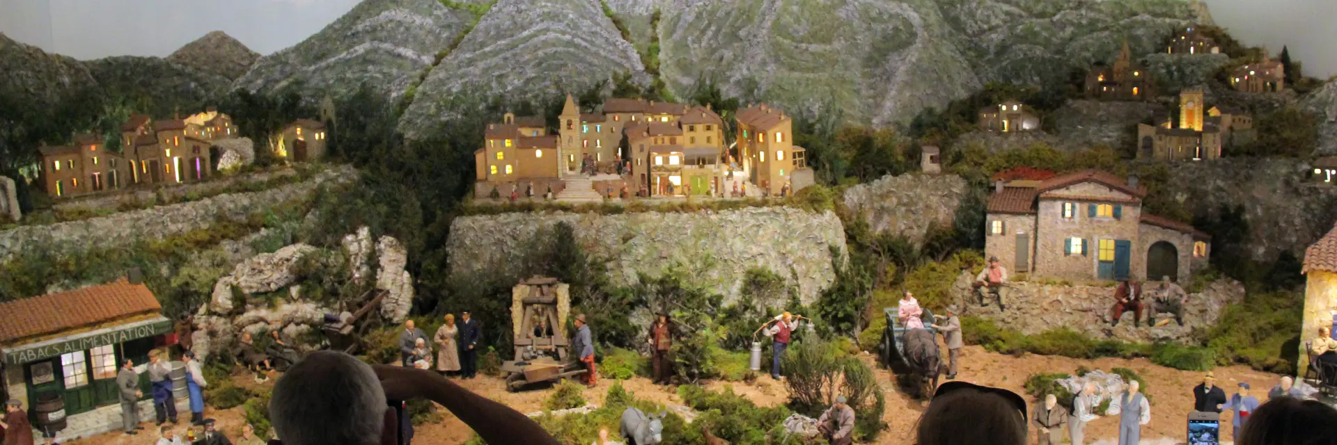 Diorama de village provençal avec maisons miniatures, collines rocheuses et personnages, observé par des visiteurs en intérieur au Petit Monde de Marcel Pagnol à Aubagne