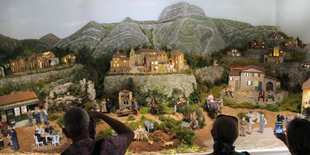 Diorama de village provençal avec maisons miniatures, collines rocheuses et personnages, observé par des visiteurs en intérieur au Petit Monde de Marcel Pagnol à Aubagne