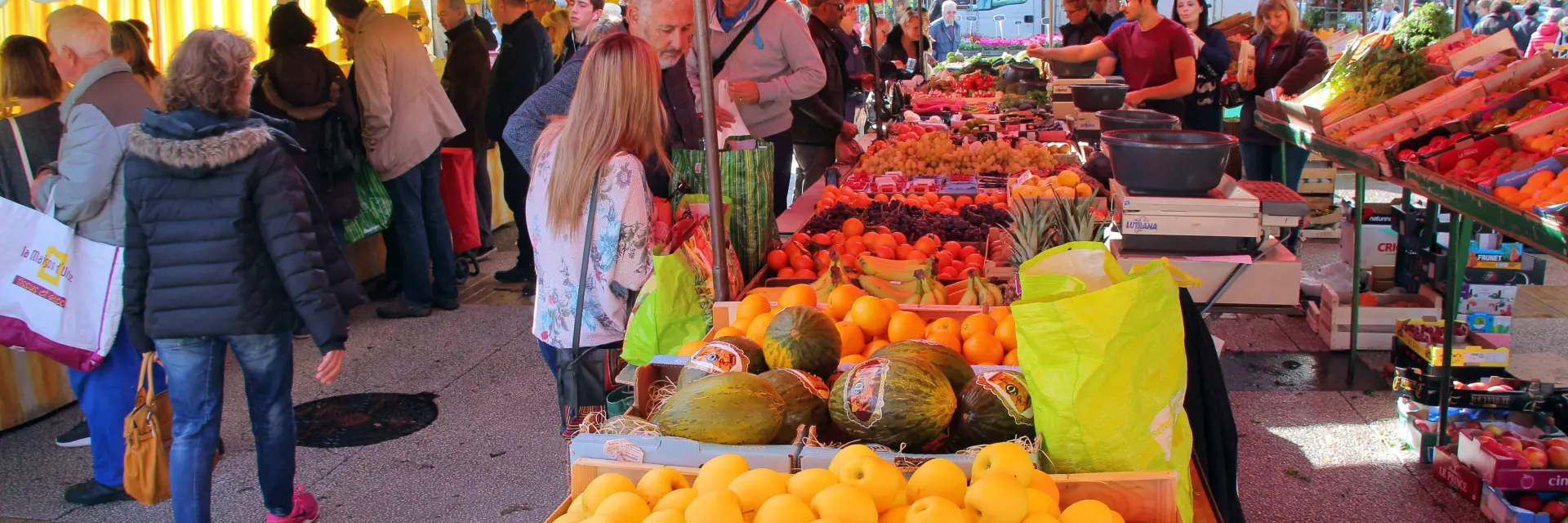 Étals colorés de fruits et légumes dans un marché en plein air, avec des visiteurs déambulant sous des auvents rouges.