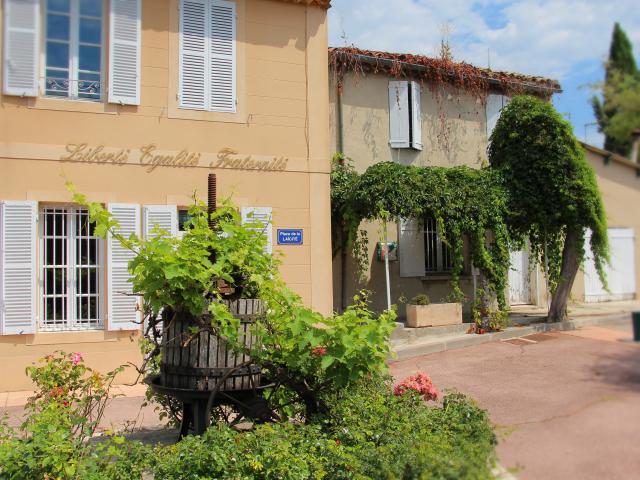 Façade de la mairie de Belcodène avec volets blancs, vigne grimpante et ancien pressoir en bois sur une petite place ensoleillée.