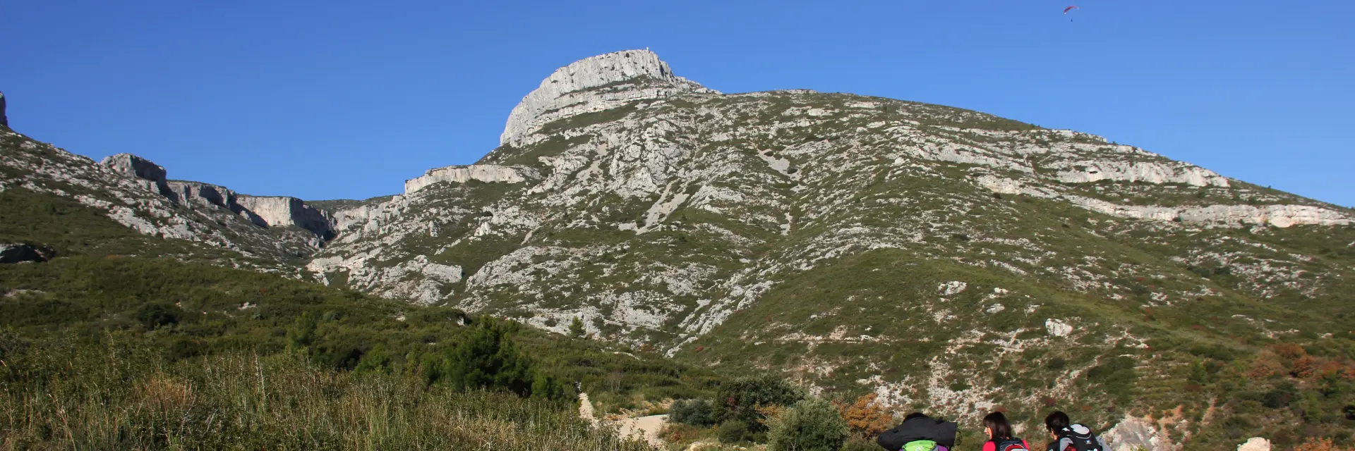 Groupe de randonneurs marchant sur un sentier de terre, entourés de végétation méditerranéenne, avec le massif rocheux du Garlaban et un ciel bleu en arrière-plan