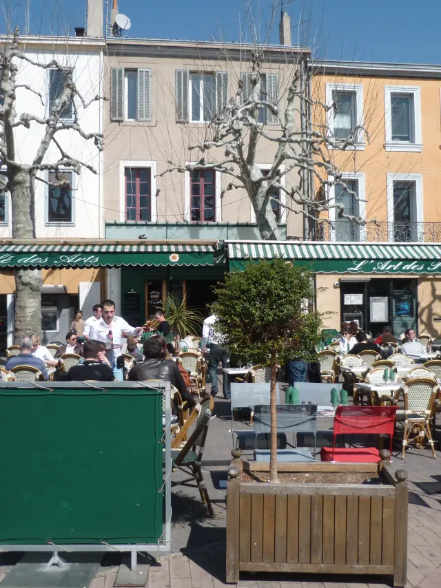 Terrasse de restaurant avec tables et chaises colorées sur une place de village au Café des Arts à Aubagne, clients attablés, façades d’immeubles et arbres taillés en arrière-plan. Café