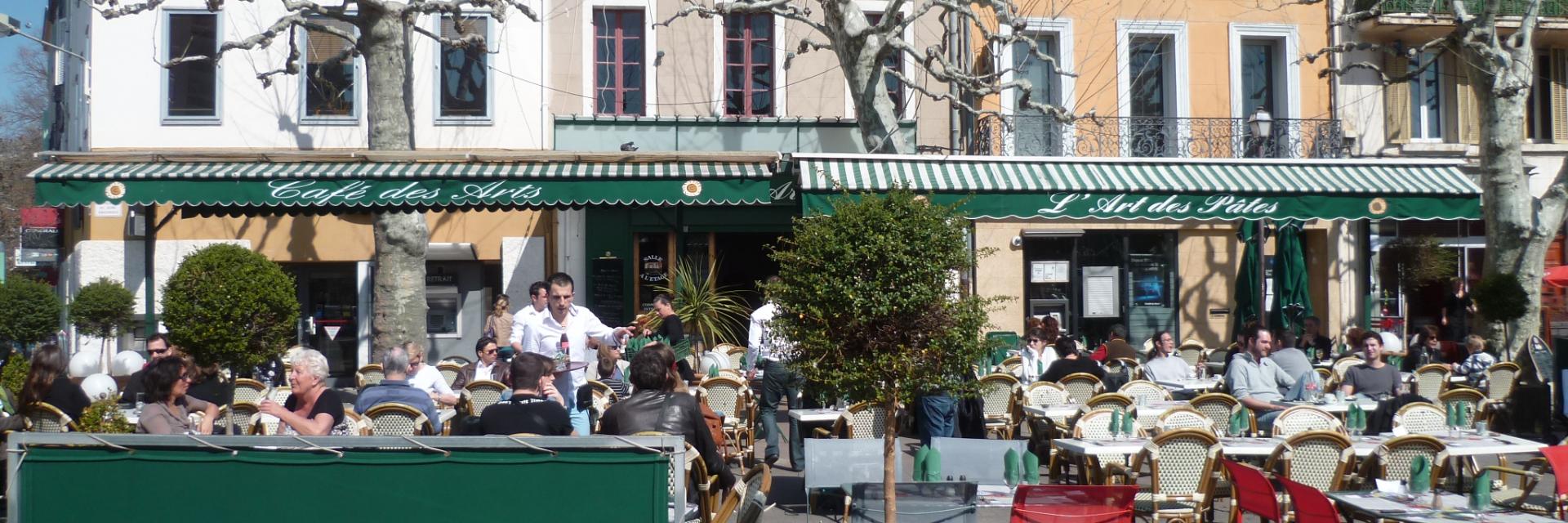 Terrasse de restaurant avec tables et chaises colorées sur une place de village au Café des Arts à Aubagne, clients attablés, façades d’immeubles et arbres taillés en arrière-plan. Café