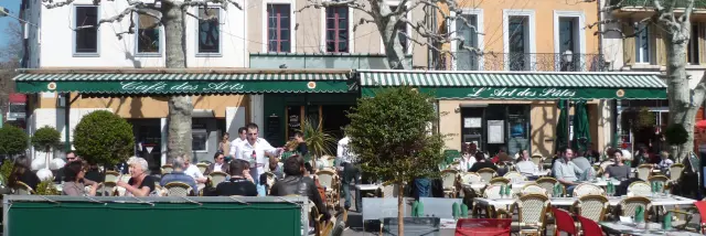 Terrasse de restaurant avec tables et chaises colorées sur une place de village au Café des Arts à Aubagne, clients attablés, façades d’immeubles et arbres taillés en arrière-plan. Café