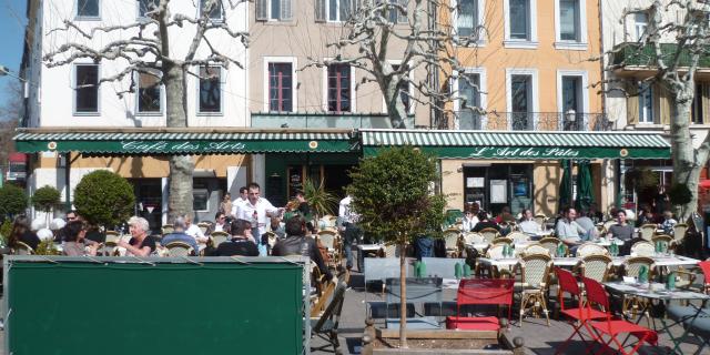 Terrasse de restaurant avec tables et chaises colorées sur une place de village au Café des Arts à Aubagne, clients attablés, façades d’immeubles et arbres taillés en arrière-plan. Café