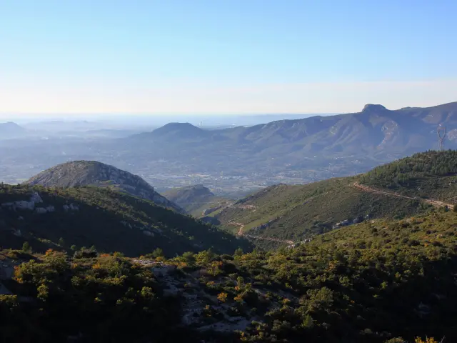 Paysage panoramique depuis une colline avec végétation de garrigue au premier plan, collines boisées au centre et Marseille à l’horizon sous un ciel bleu.