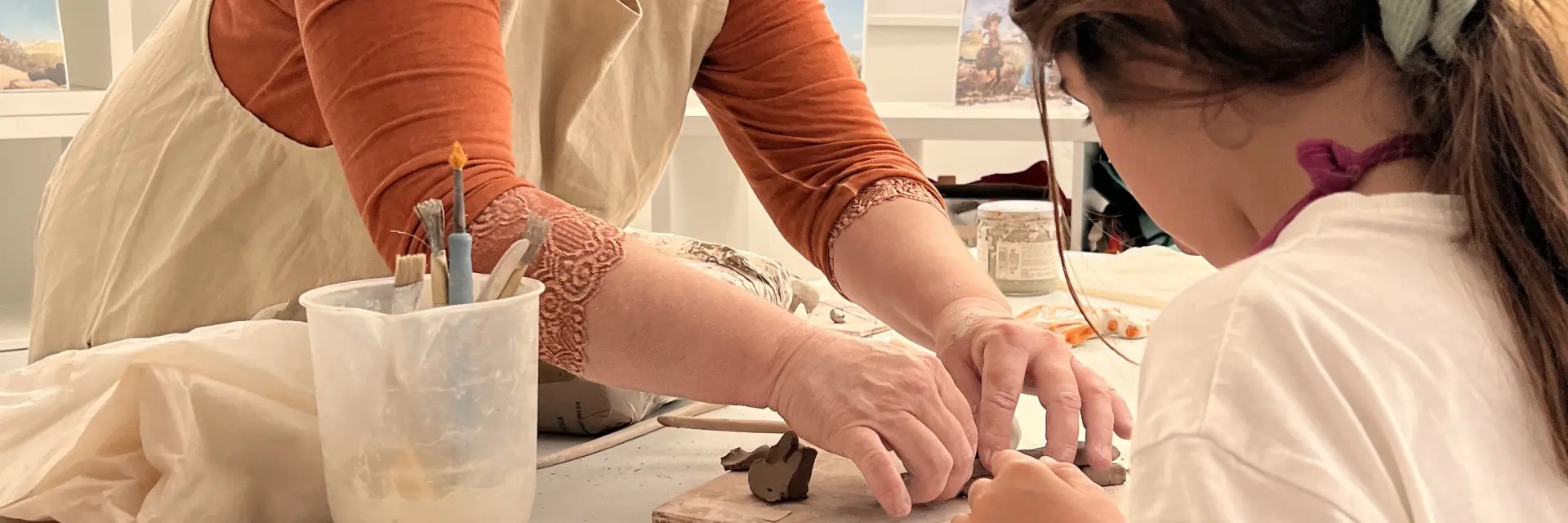 Une artisane guidant une enfant dans un atelier de modelage en argile, en façonnant une petite sculpture sur une table avec des outils de poterie.