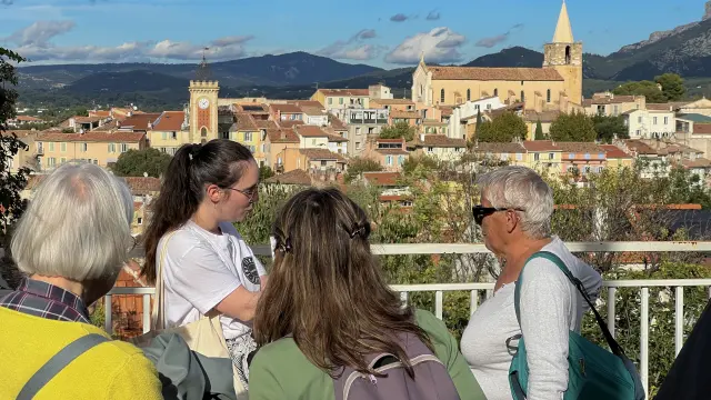 Groupe de personnes participant à une visite guidée, observant un village provençal avec église, clocher et maisons aux toits en tuiles en arrière-plan à Aubagne