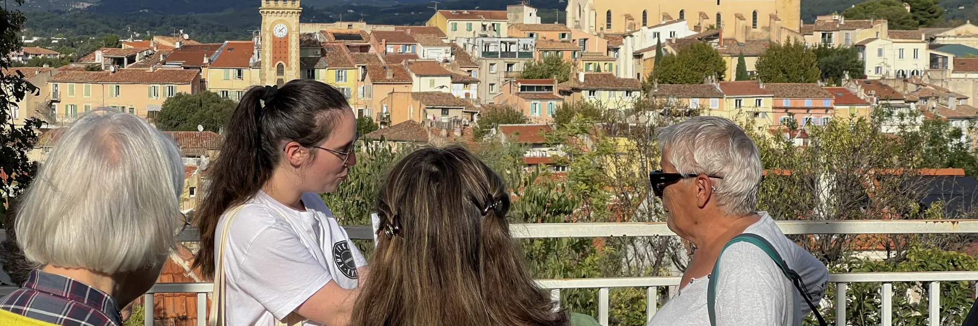 Groupe de personnes participant à une visite guidée, observant un village provençal avec église, clocher et maisons aux toits en tuiles en arrière-plan à Aubagne
