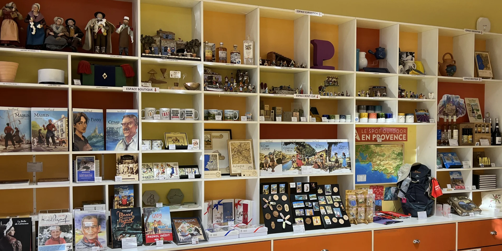 Boutique intérieure avec étagères murales remplies de souvenirs, livres, cartes postales et objets décoratifs, présentés dans un espace lumineux à l'Office de Tourisme d'Aubagne
