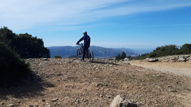 Cycliste en VTT à l'arrêt sur un sentier rocailleux, observant le panorama vers Aubagne et les montagnes aux alentours sous un ciel bleu.