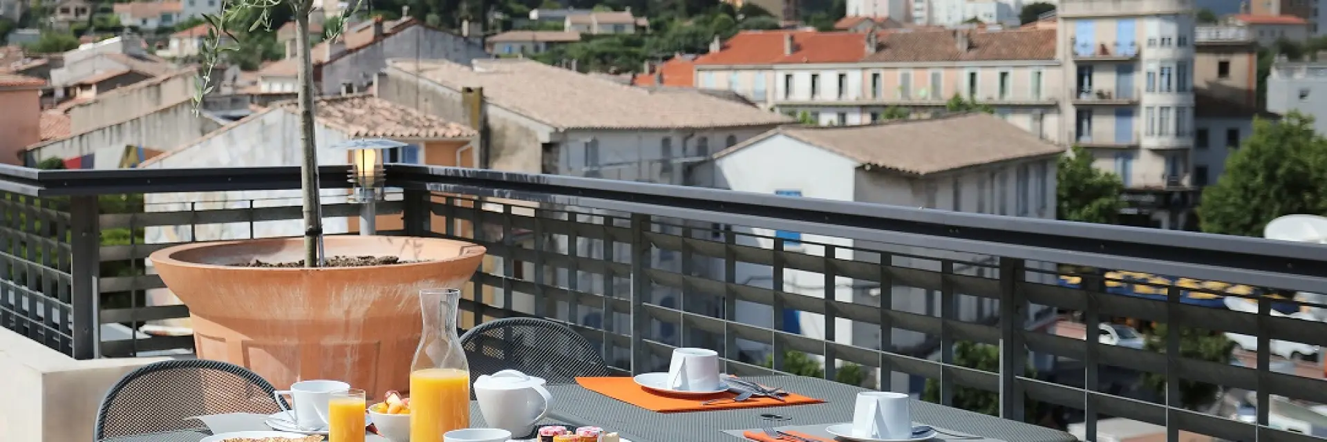 Table de petit-déjeuner dressée sur une terrasse d’hôtel avec jus d’orange, café et viennoiseries, offrant une vue dégagée sur les toits de la ville et les collines environnantes