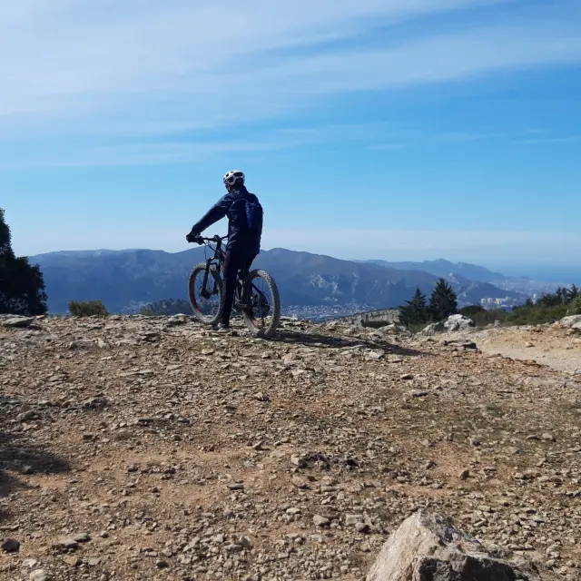 Cycliste en VTT à l'arrêt sur un sentier rocailleux, observant le panorama vers Aubagne et les montagnes aux alentours sous un ciel bleu.
