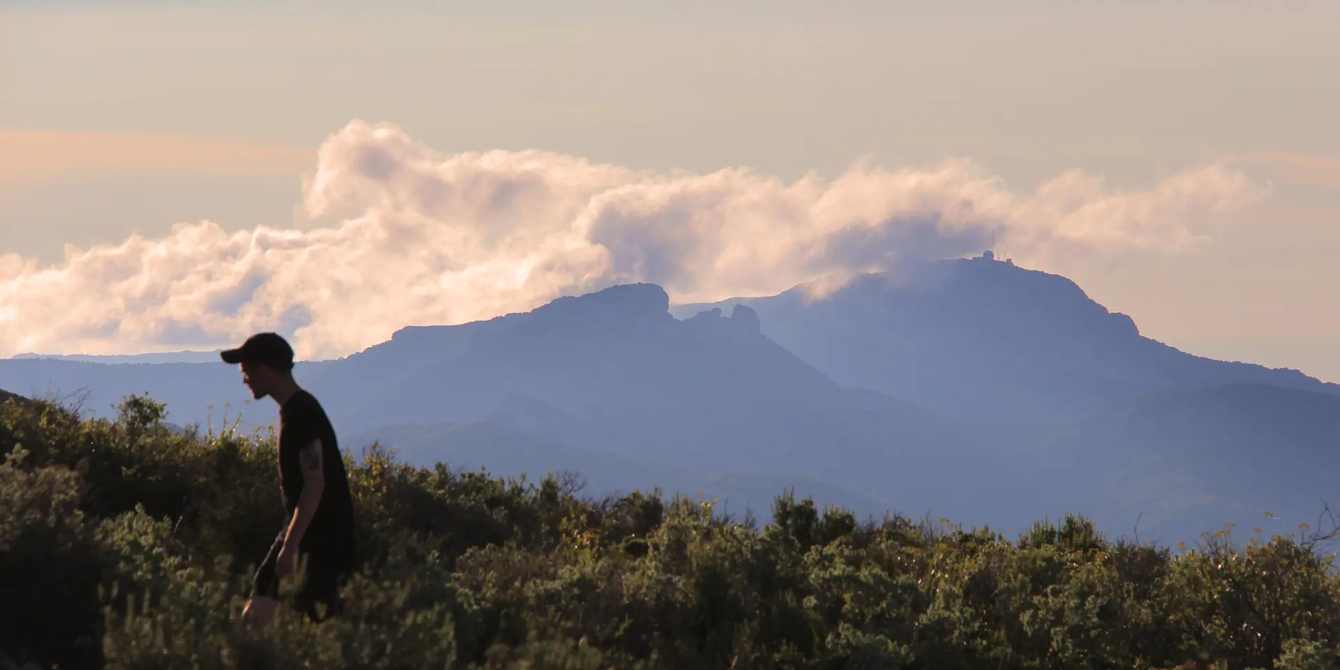 Randonneur marchant dans la garrigue du massif du Garlaban avec vue sur les reliefs montagneux et les nuages en Provence.