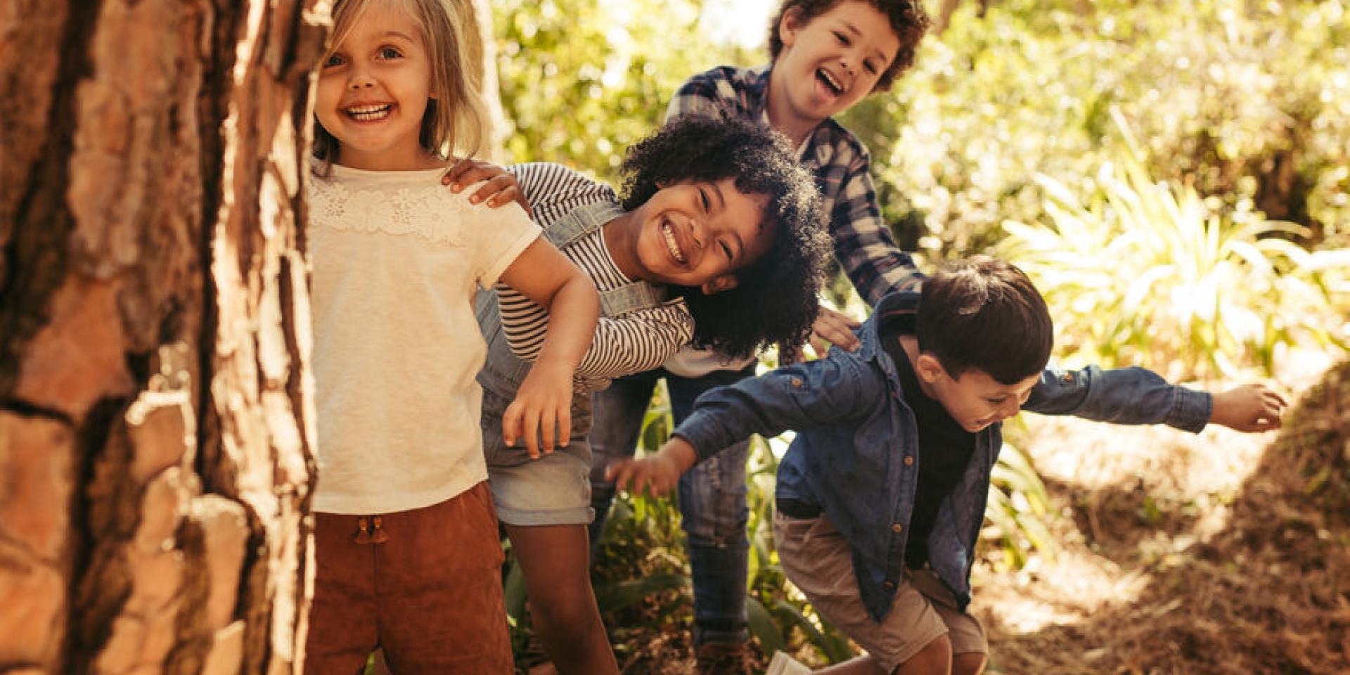 Cute smiling kids peeking out from behind the tree in the park. Group of children enjoying playing hide and seek in a forest.