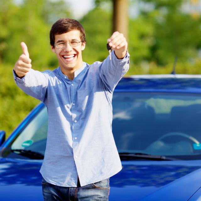 Young happy man in glasses standing in front of his new car and showing thumb up and car key