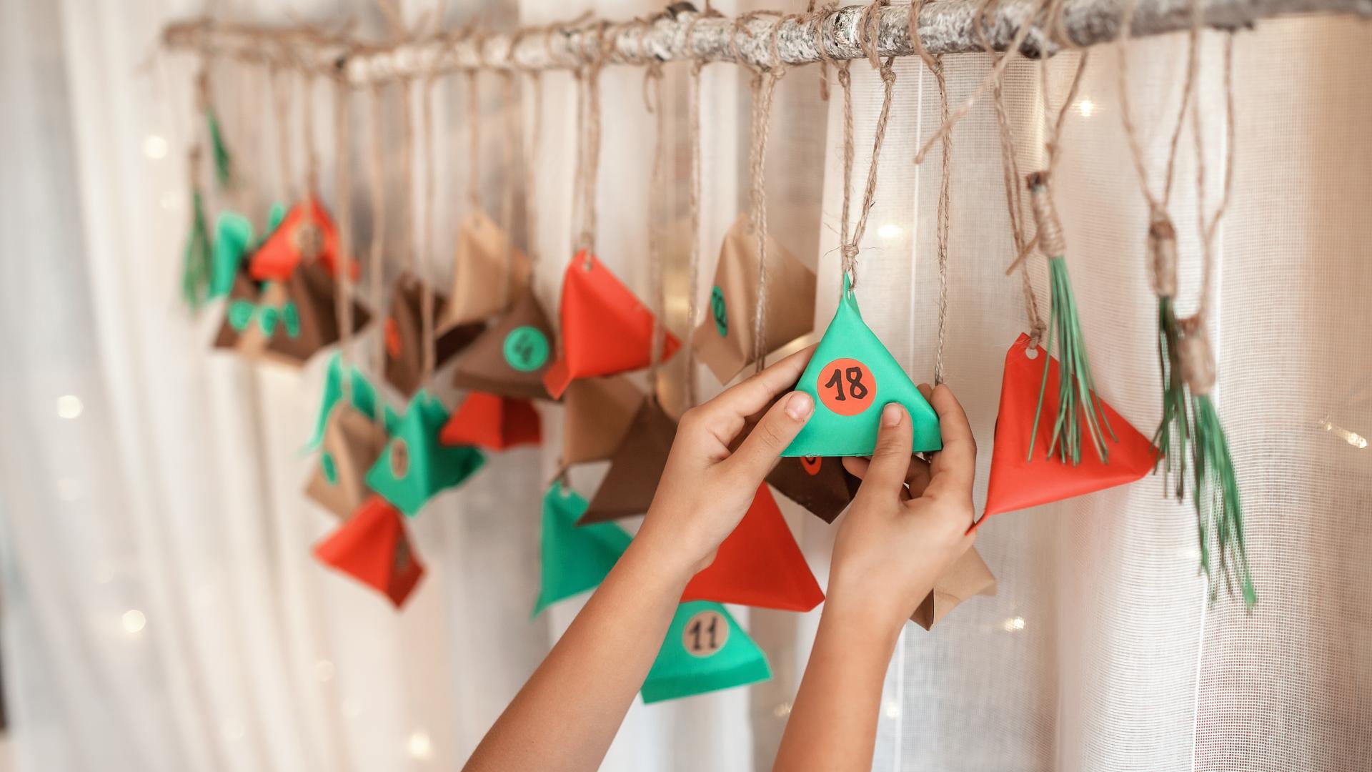 Cute little kid opening handmade advent calendar with color paper triangles. Sweets and candies are hidden in colorful triangles hanging on branch. Seasonal activity for kids, family winter holidays