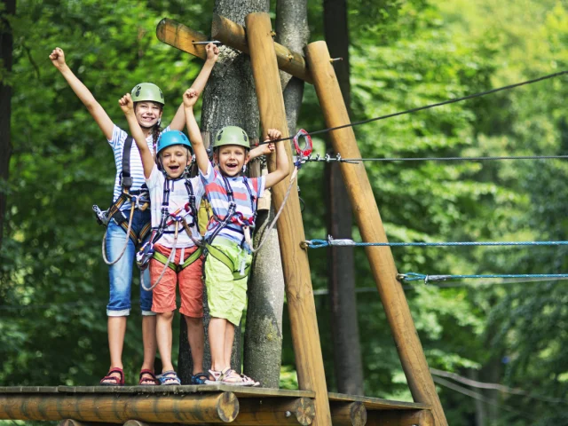 10 year old girl with her 7 year old brothers, wearing helmets, on a wooden platform holding zip line in the adventure park. The children smile at the camera and applaud.