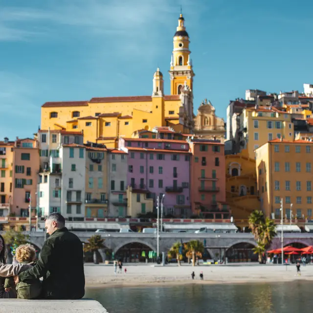 Famille en promenade hivernale à Menton face aux façades colorées de la vieille ville, sur la Côte d’Azur