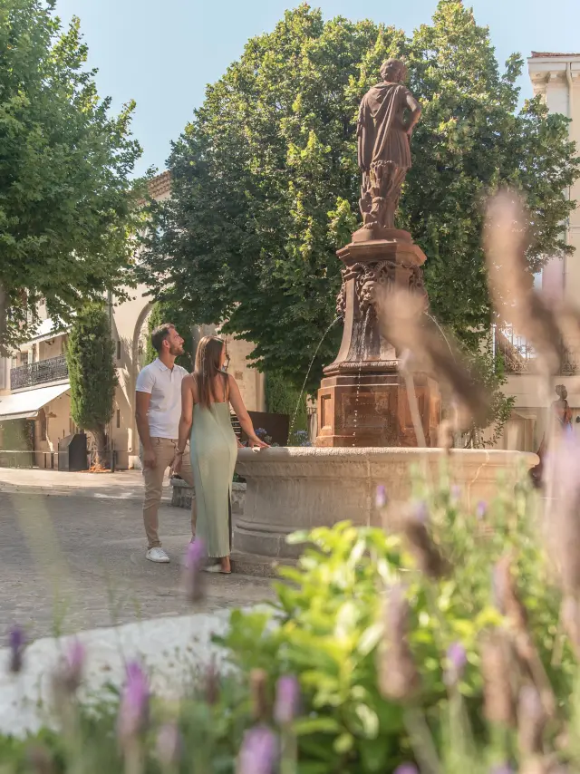 Un couple admire la fontaine centrale de la place Lamy à Mougins, en été, entouré de lavande et de bâtisses provençales