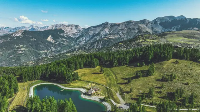Vue aérienne de la station de Valberg avec son lac artificiel entouré de forêts et de montagnes alpines, sous un ciel d'été bleu.