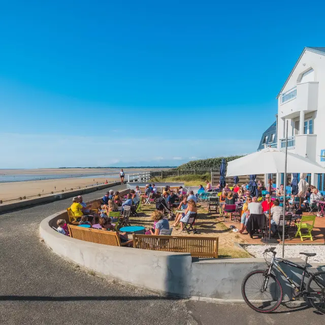 Terrasse de café bondée en bord de plage avec des gens assis et des vélos garés