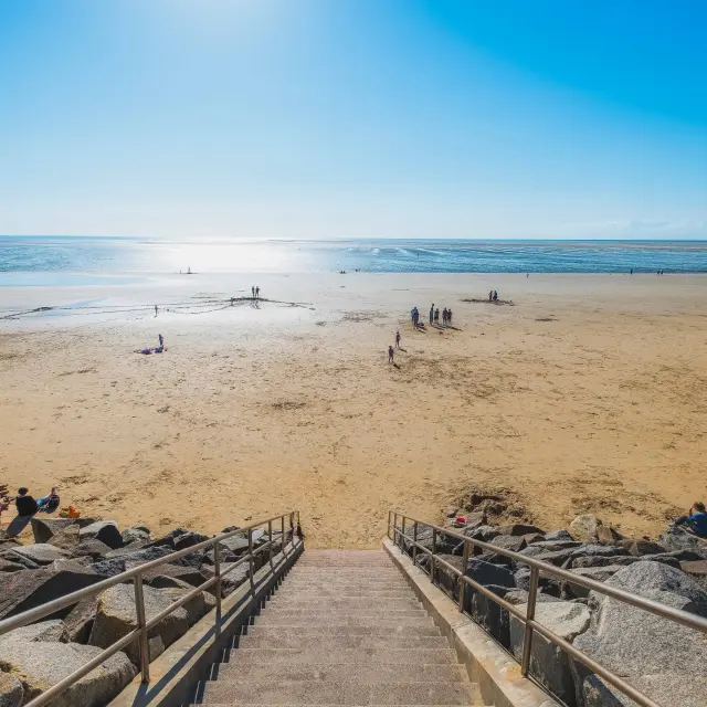 Plage avec des personnes se baignant, des parasols et des transats sur le sable, bordée par l'océan