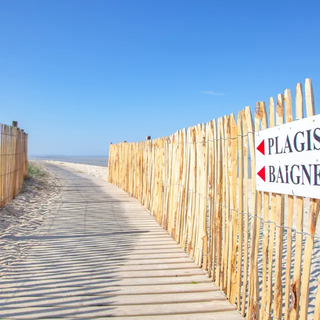 Passerelle en bois menant à une plage avec un panneau indiquant Plagistes Baigneurs