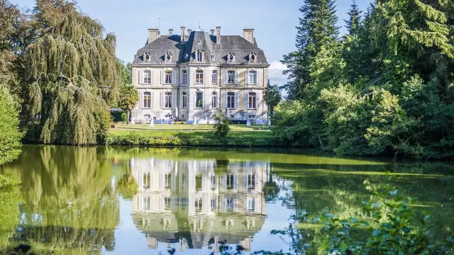 Manoir à deux étages avec toit en ardoise, reflété dans un lac entouré d'arbres