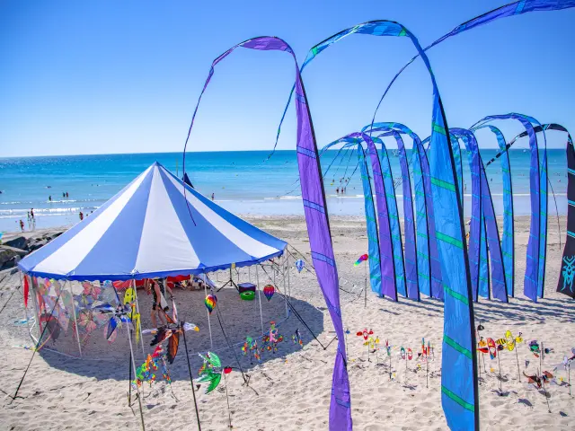 Bunte Sonnenschirme in einer Reihe auf einem weißen Sandstrand mit vertäuten Segelbooten im Hintergrund