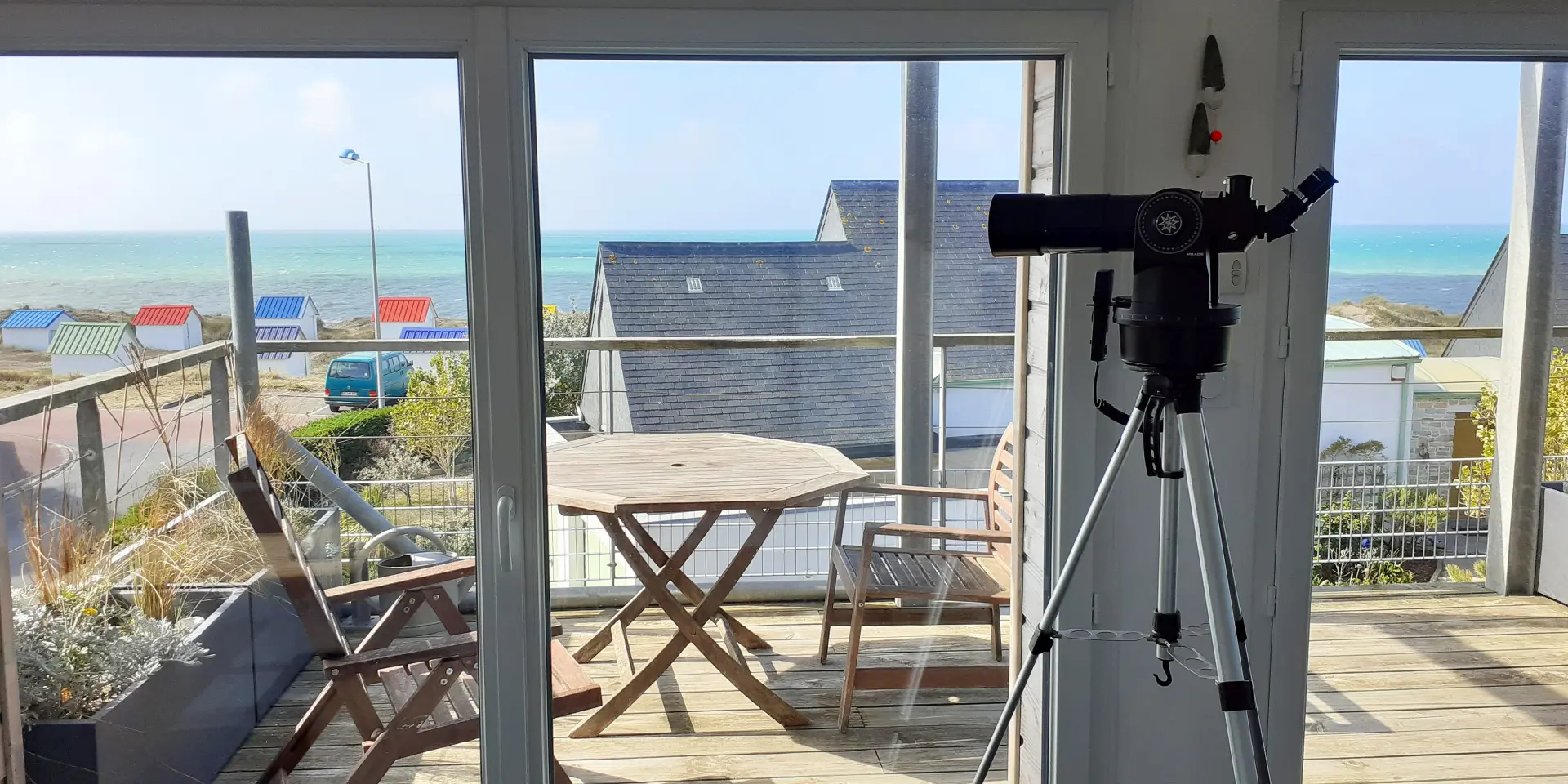 Terrasse de café en bord de mer avec des tables, chaises et parasols, vue sur l’eau et des voiliers amarrés