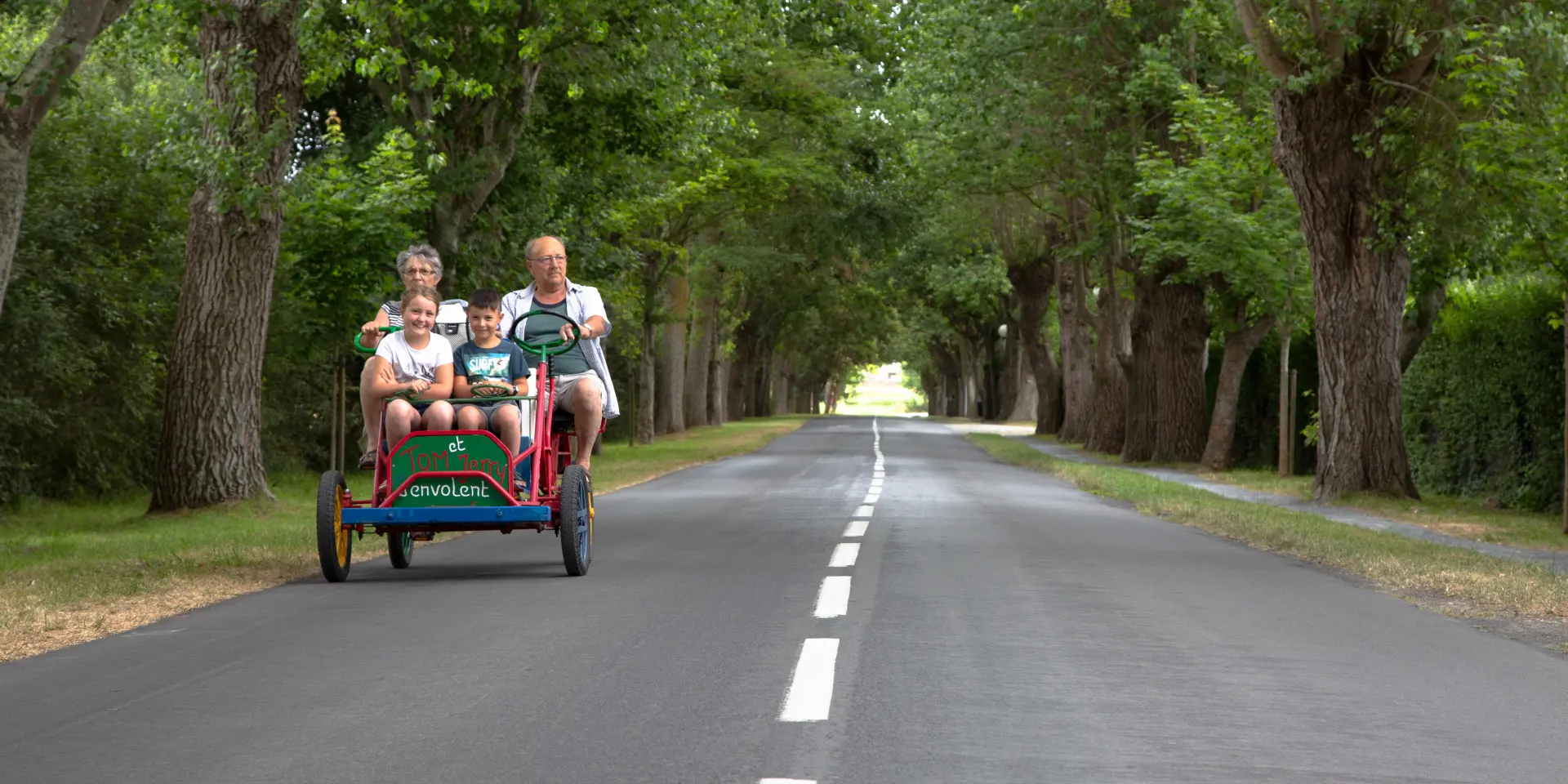 Une famille de quatre personnes sur une rosalie à quatre places sur une route bordée d'arbres à Hauteville sur Mer