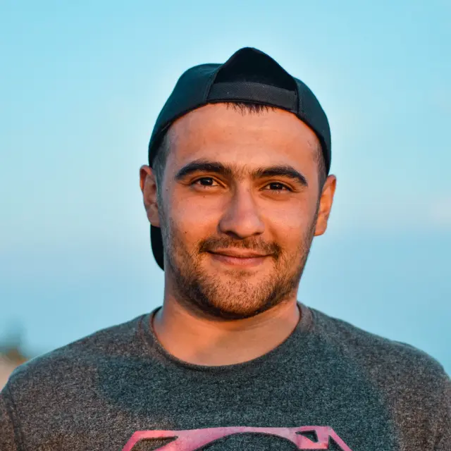 Homme avec barbe et casquette noire, vêtu d’un pull à col rond, souriant face à l’appareil photo