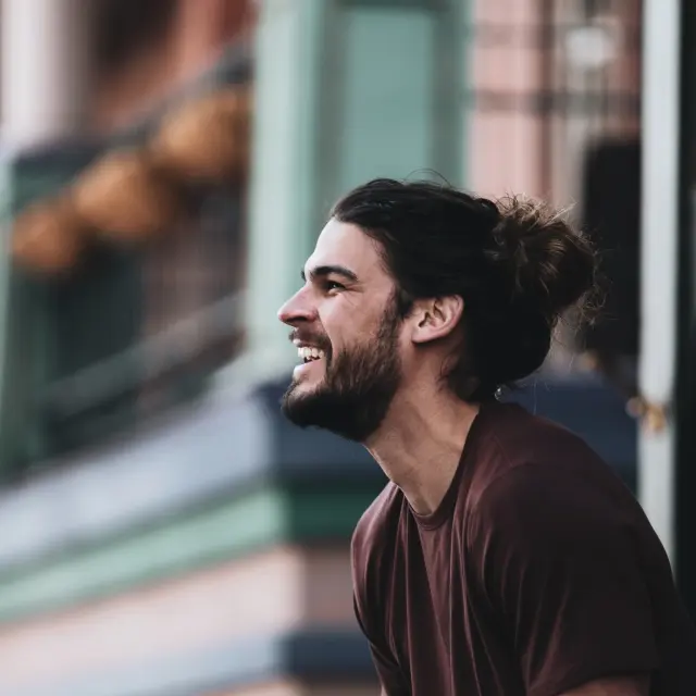 Un homme avec une barbe et un bun, portant un t-shirt marron, rit en marchant dans une rue urbaine.