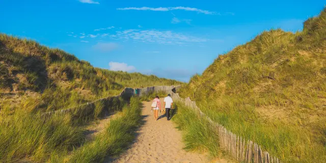 Zwei Erwachsene und ein Kind gehen auf einem Sandweg zwischen grasbewachsenen Dünen