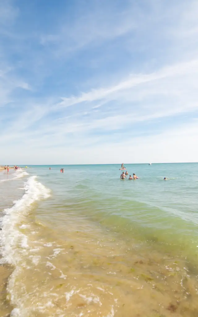 Plage avec des personnes nageant et des voiliers amarrés au large, ciel bleu avec quelques nuages