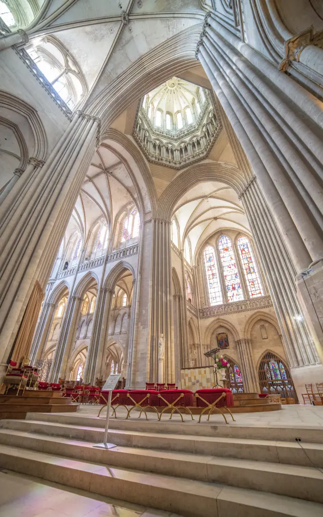Central nave of a Gothic cathedral with ribbed vaults, stained glass windows, and rows of red pews