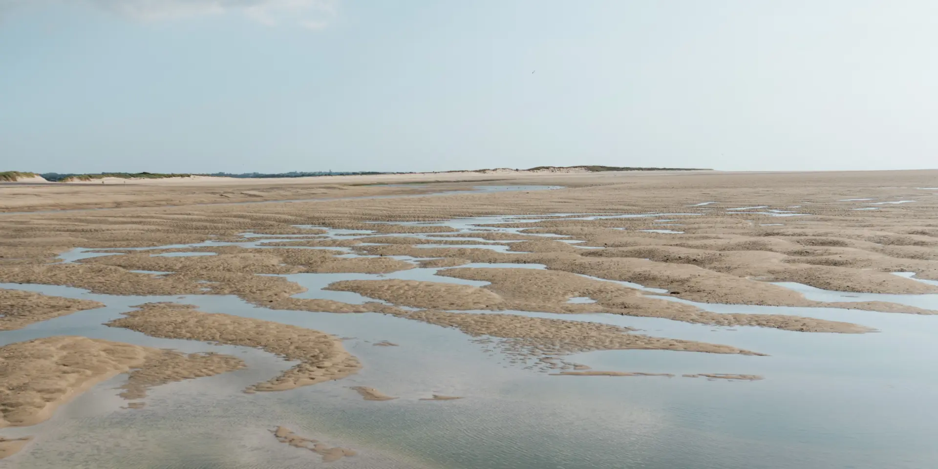 Bord de mer avec sable humide, flaques d’eau et traces de pas, sous un ciel nuageux