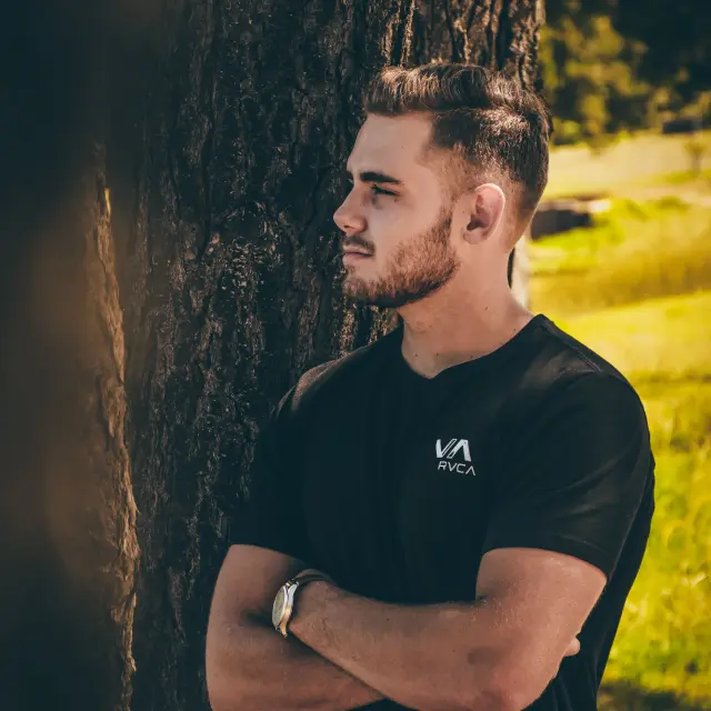 A man with a black t-shirt and a wristwatch, standing near a tree