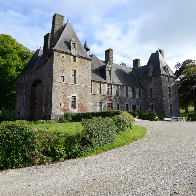 Manoir en pierre avec tourelles et toits pointus, entouré d'une allée gravillonnée et de haies taillées