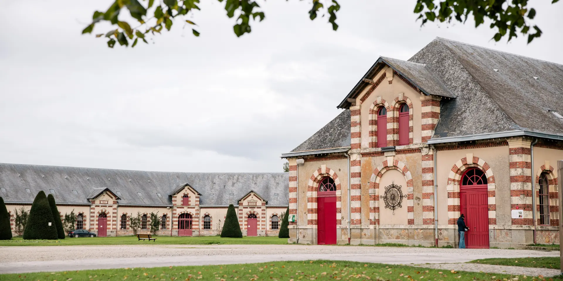 Église en brique rouge avec deux portes rouges et des vitraux, entourée d’un jardin