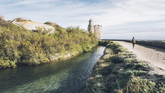 Phare en pierre sur une falaise, avec une personne marchant sur un chemin de terre au bord de l’eau