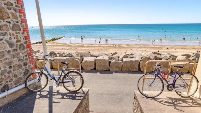 Deux vélos attachés à un poteau près d’une plage avec des voiliers amarrés et des bâtiments en arrière-plan
